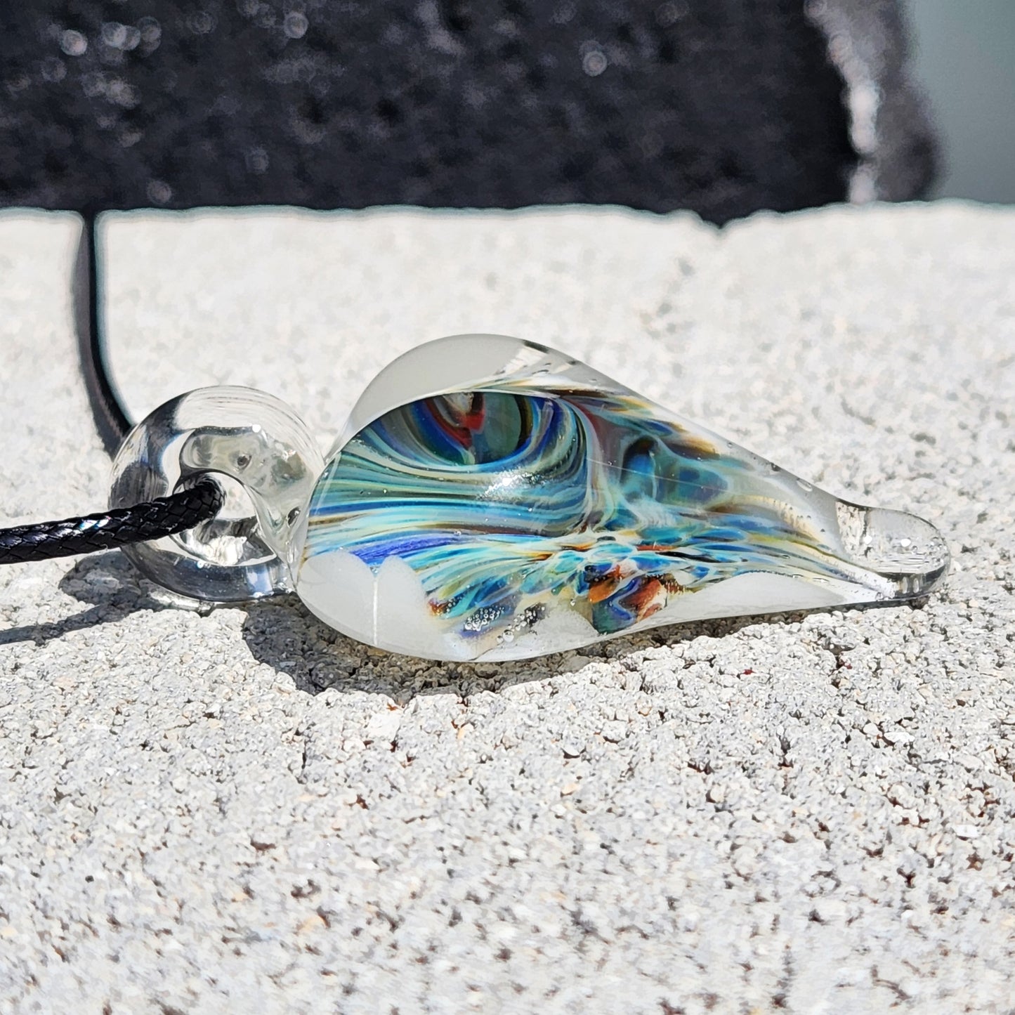 Side profile of handmade blown glass heart pendant with swirling blue and green design, lying flat on a textured stone surface.