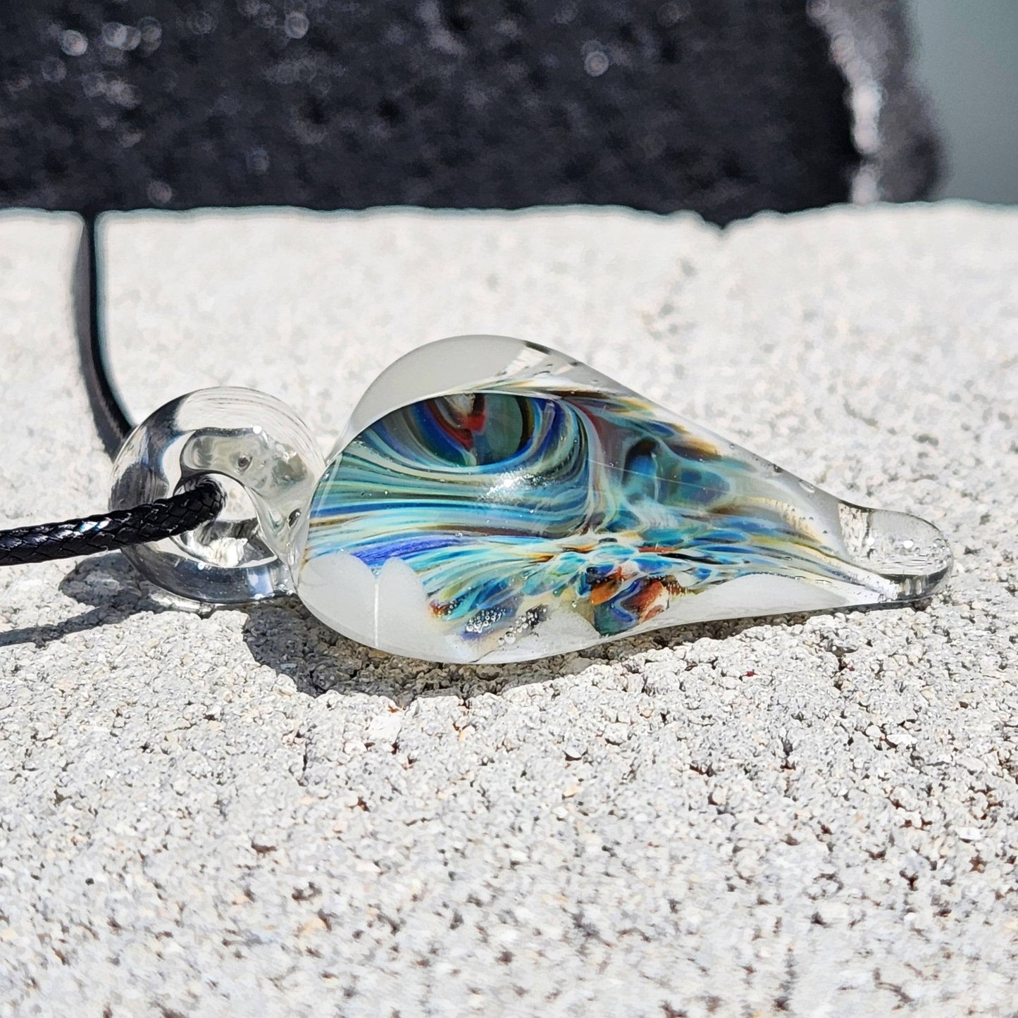 Side profile of handmade blown glass heart pendant with swirling blue and green design, lying flat on a textured stone surface.