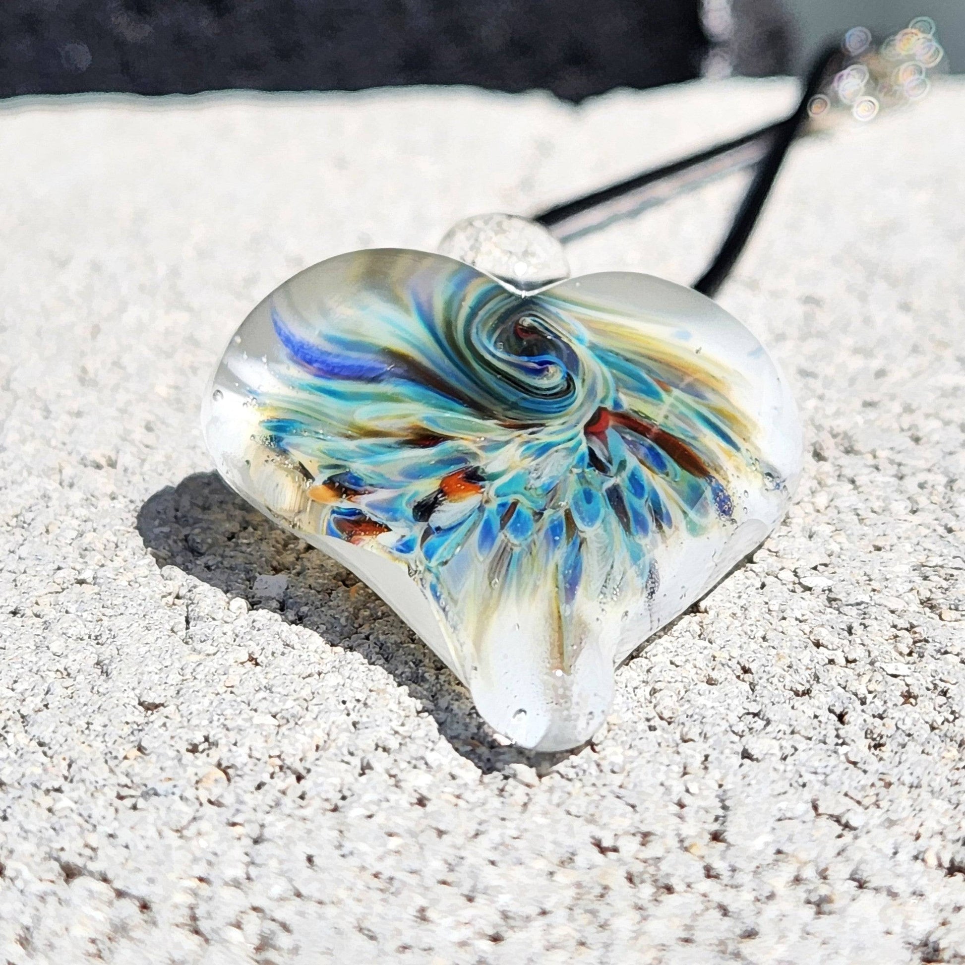 Heart-shaped handmade glass pendant featuring a vibrant implosion design in blue and green tones, resting on a textured stone surface.