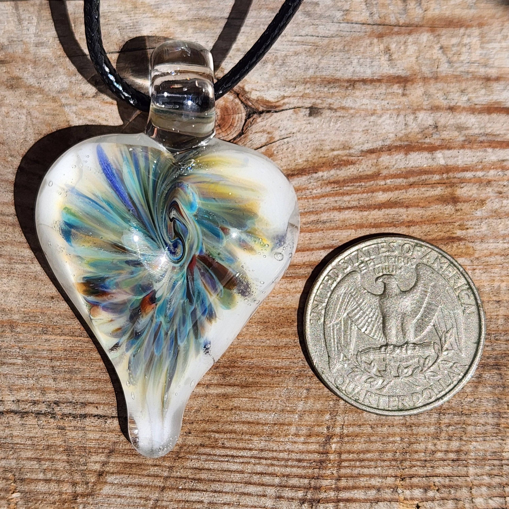 Heart-shaped handmade blown glass pendant with trippy swirl design next to a US quarter for size comparison on wooden surface.
