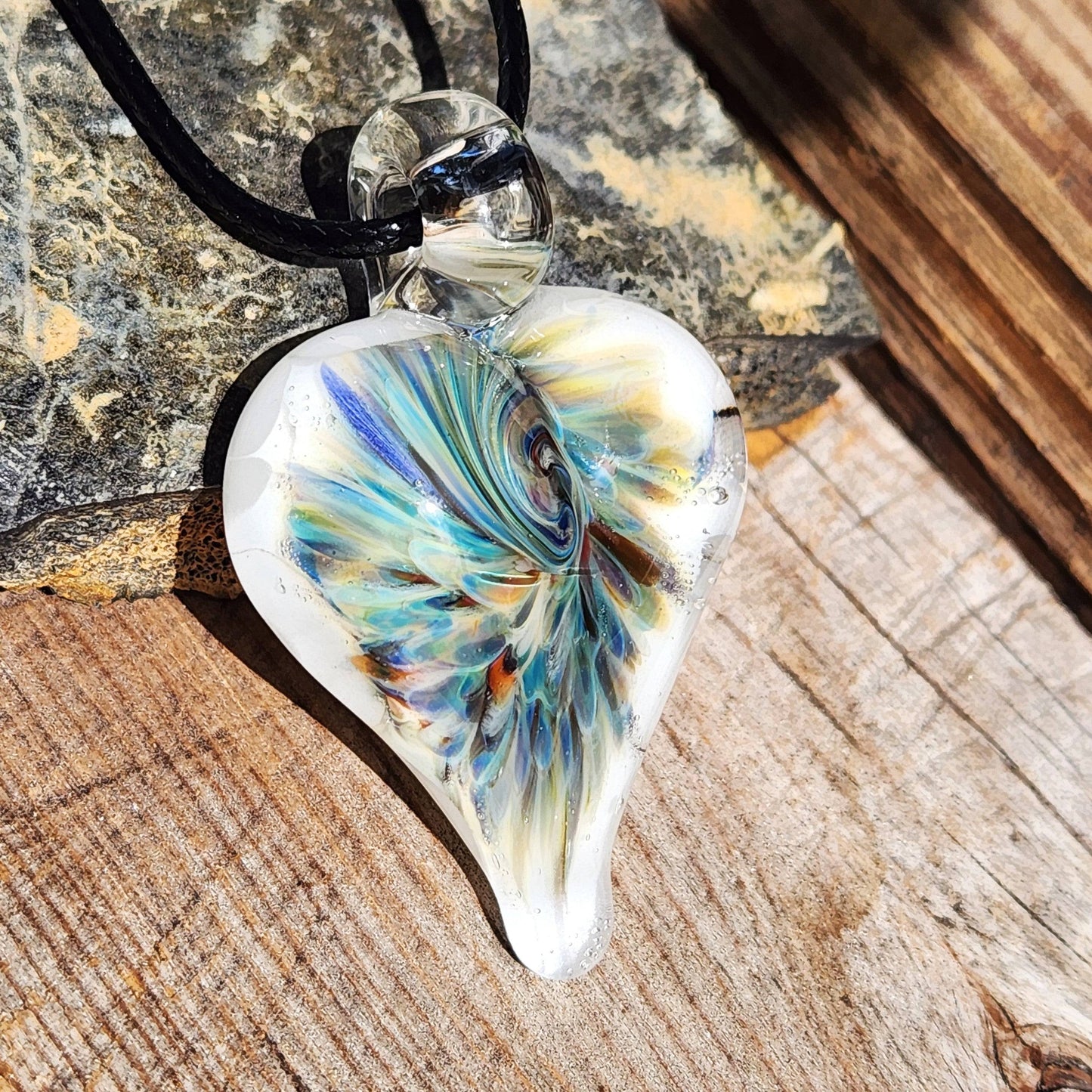 Handcrafted heart-shaped glass pendant with vibrant trippy design in blue and green, displayed on rustic wood with natural lighting.