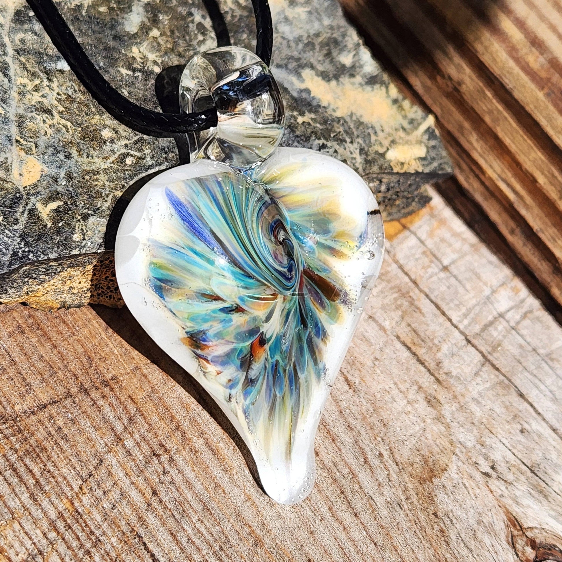 Handcrafted heart-shaped glass pendant with vibrant trippy design in blue and green, displayed on rustic wood with natural lighting.