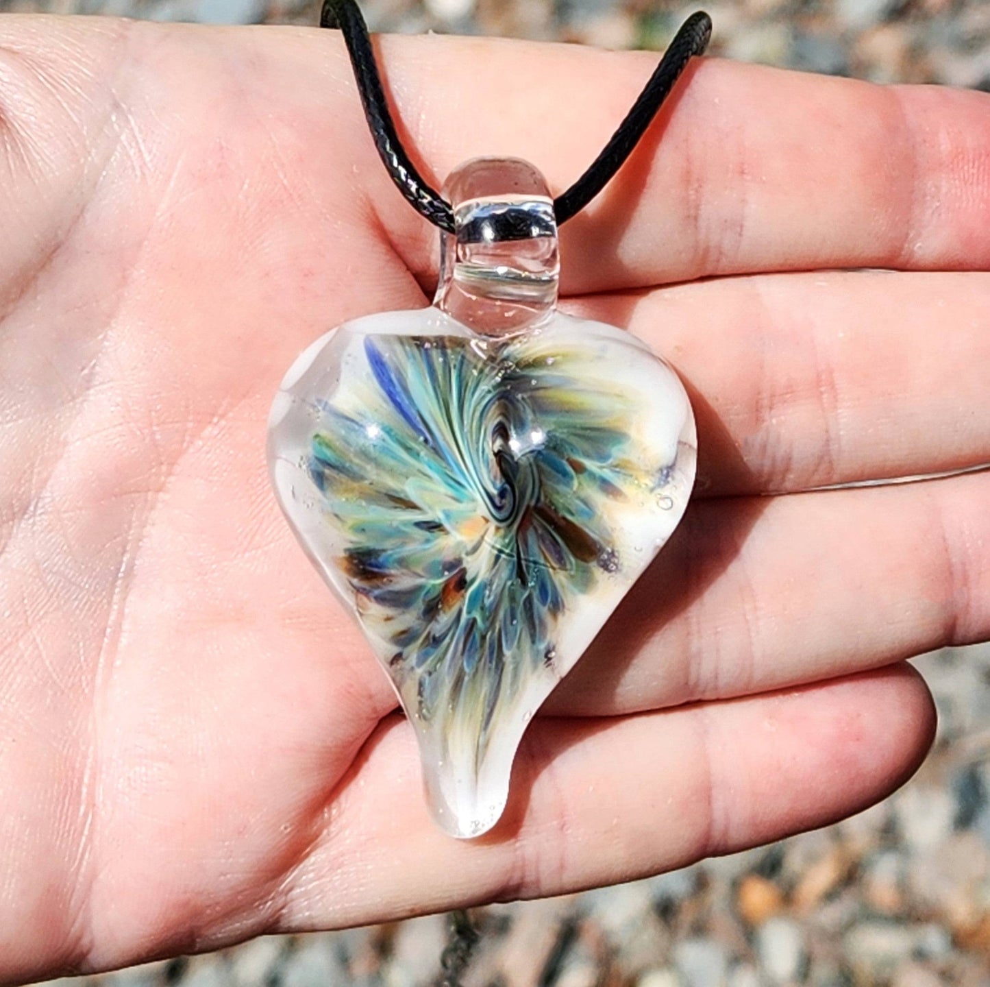Hand holding a handmade heart-shaped glass pendant with swirling trippy design in blue, green, and white, shown in natural sunlight.
