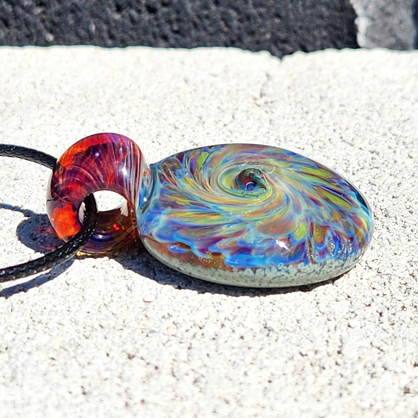 One of a kind handmade glass pendant with colorful spiral pattern and red loop, lying flat on textured stone surface.