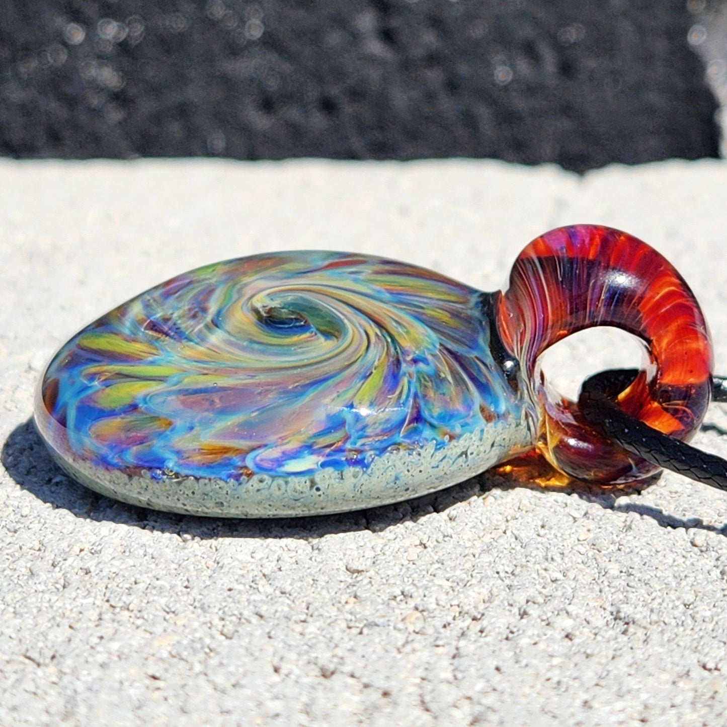 Artisan glass pendant necklace with swirling rainbow pattern and red loop, shown on stone surface with natural light.