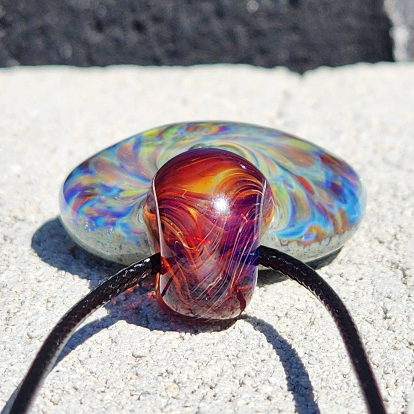 Close-up of red-amber loop on handmade glass pendant necklace with swirling multicolor design, shown on stone.