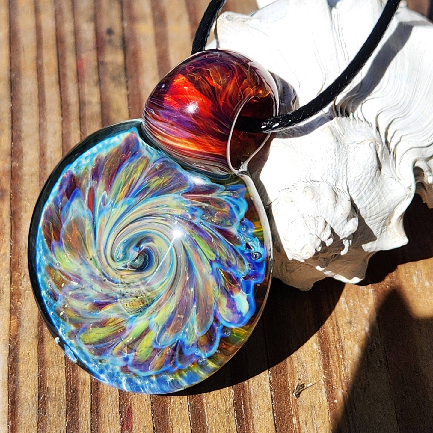 Handmade blown glass pendant necklace featuring vivid blue, purple, and red swirls, displayed on wood with natural lighting.