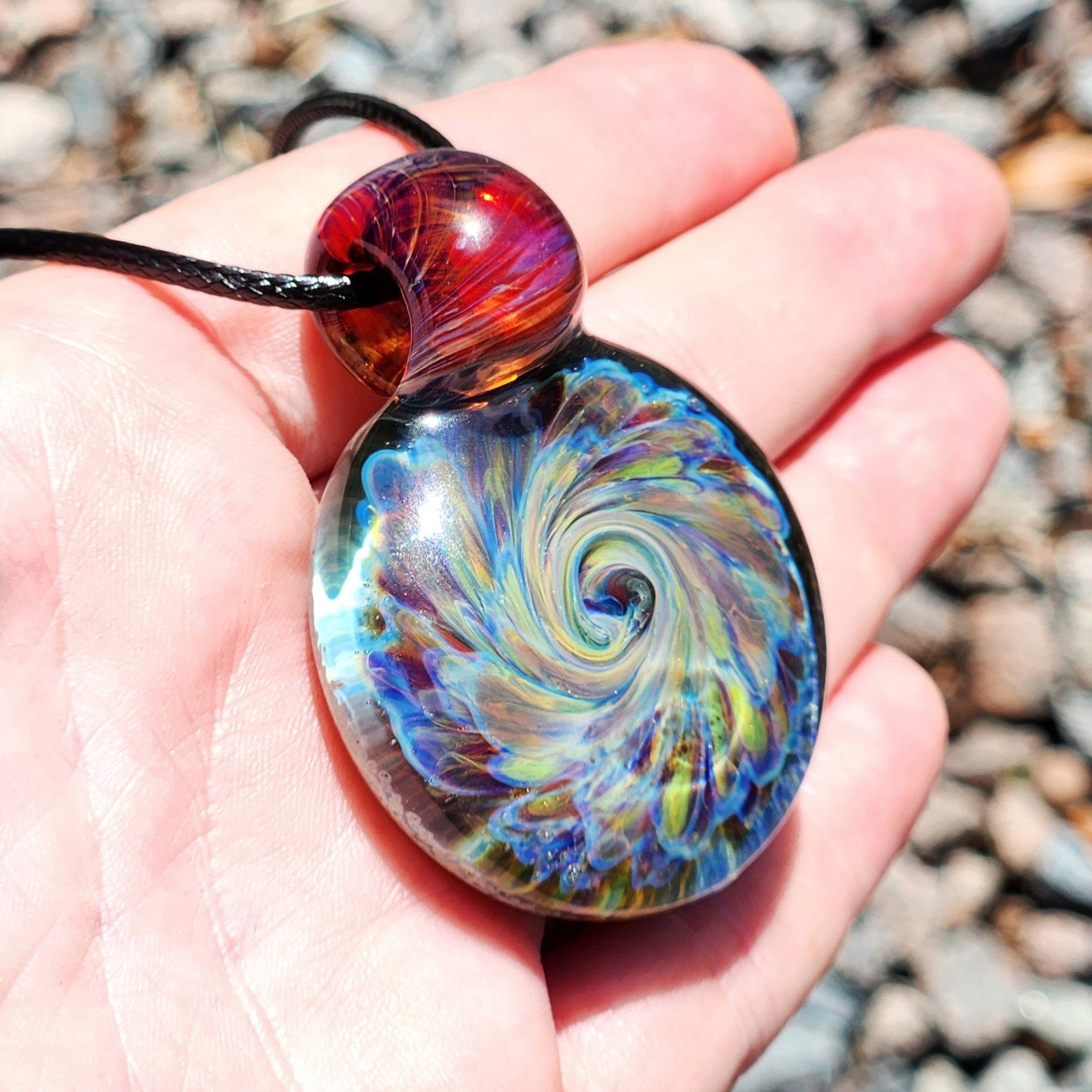 Hand holding handmade glass pendant necklace with swirling rainbow colors and red loop, one of a kind jewelry.