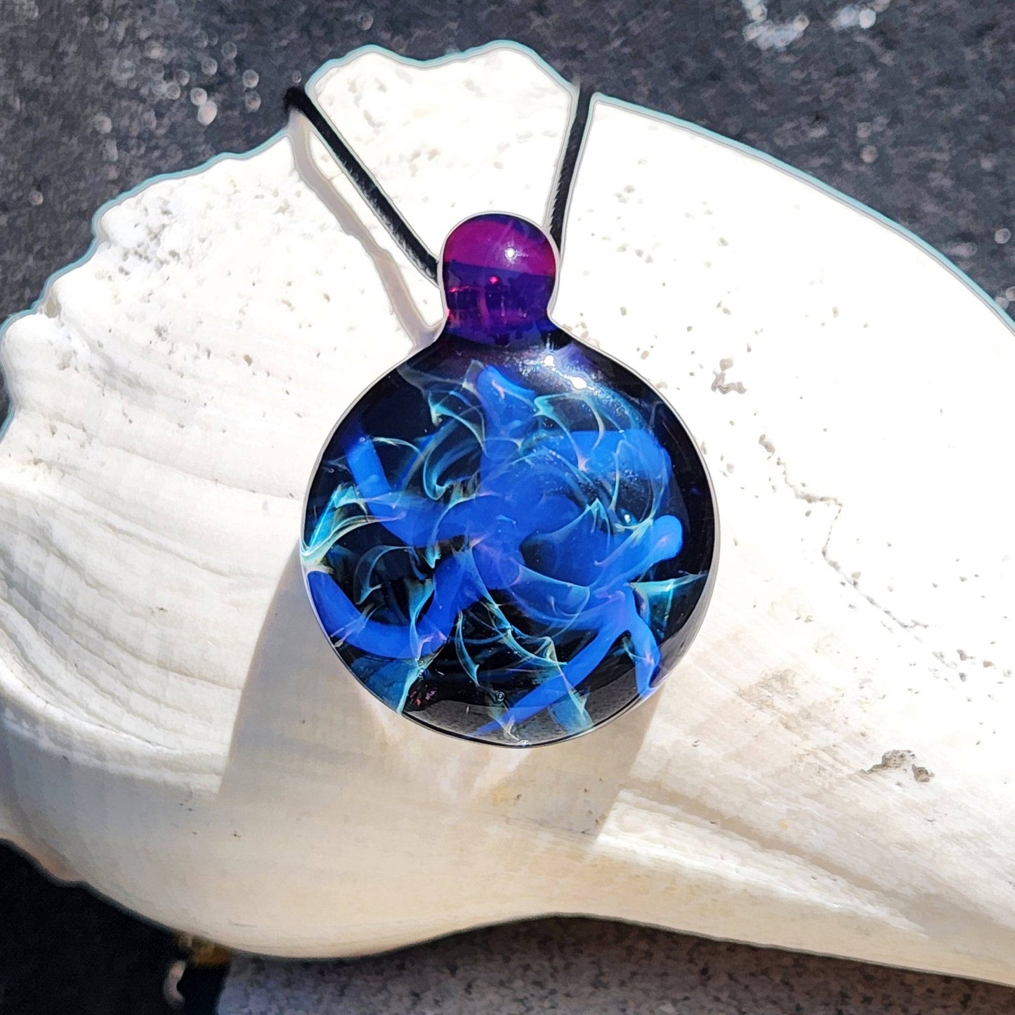 Handmade Blue Swirl Glass Pendant Necklace with purple loop and black background, displayed on a white seashell in sunlight.