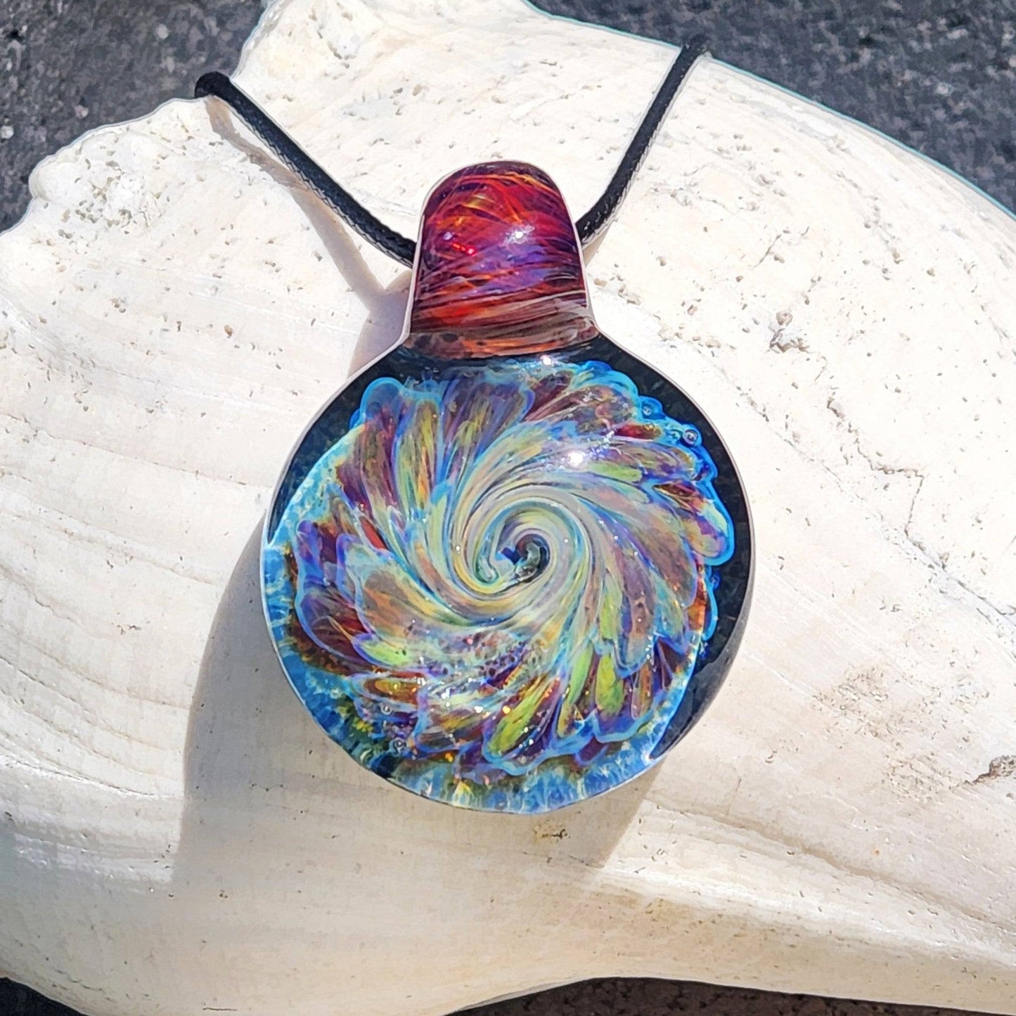 Multicolor glass pendant with swirling rainbow design and red-amber loop, handmade necklace in sunlight on white seashell.