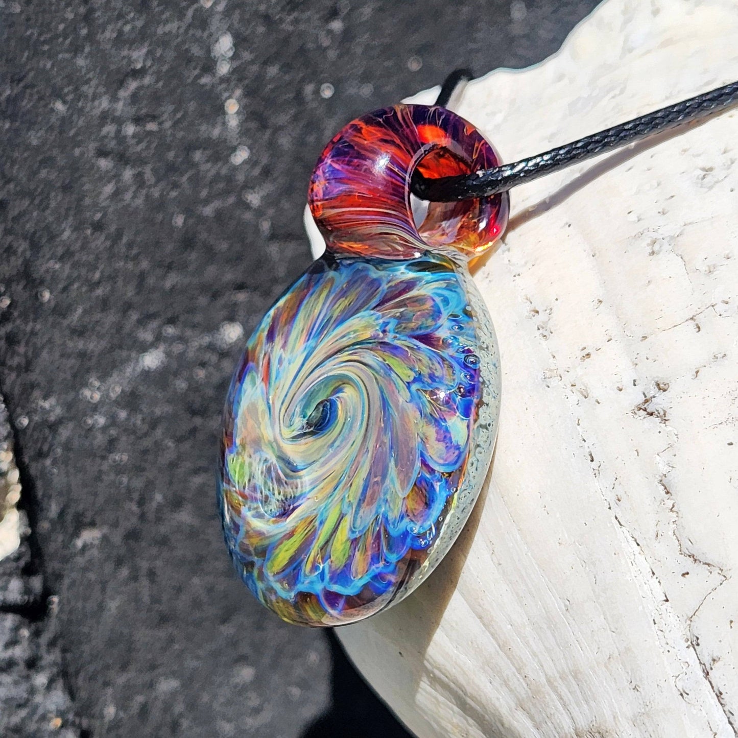 Swirling rainbow glass pendant with red loop, handmade necklace resting on white shell, vibrant colors in sunlight.