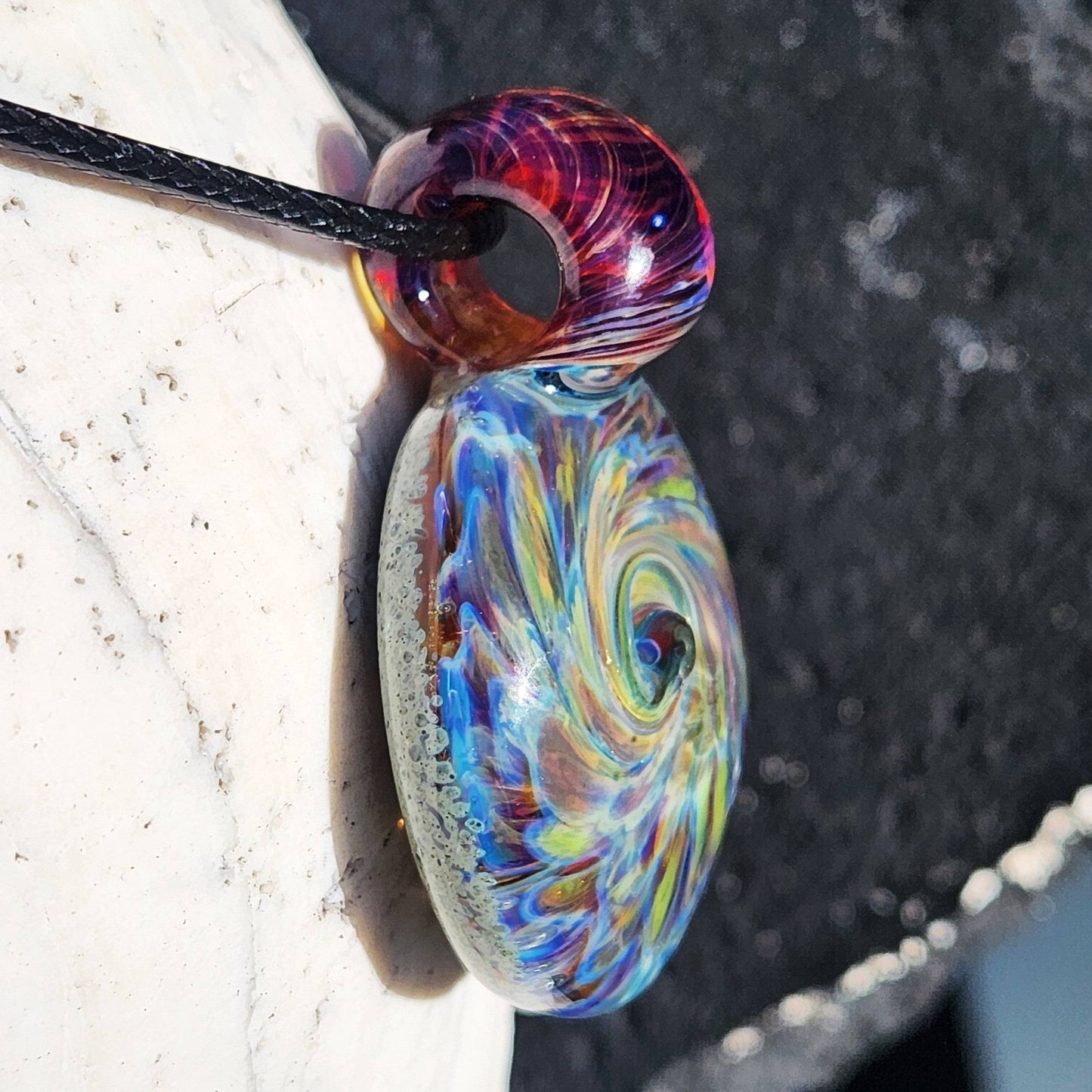 Side angle of multicolor blown glass pendant with swirling design and red-amber loop, handmade necklace on shell.