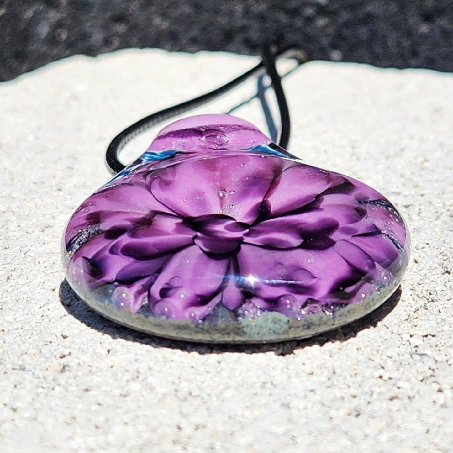 Front view of purple flower glass pendant on textured surface, showcasing intricate petal design. Handcrafted jewelry gift idea.