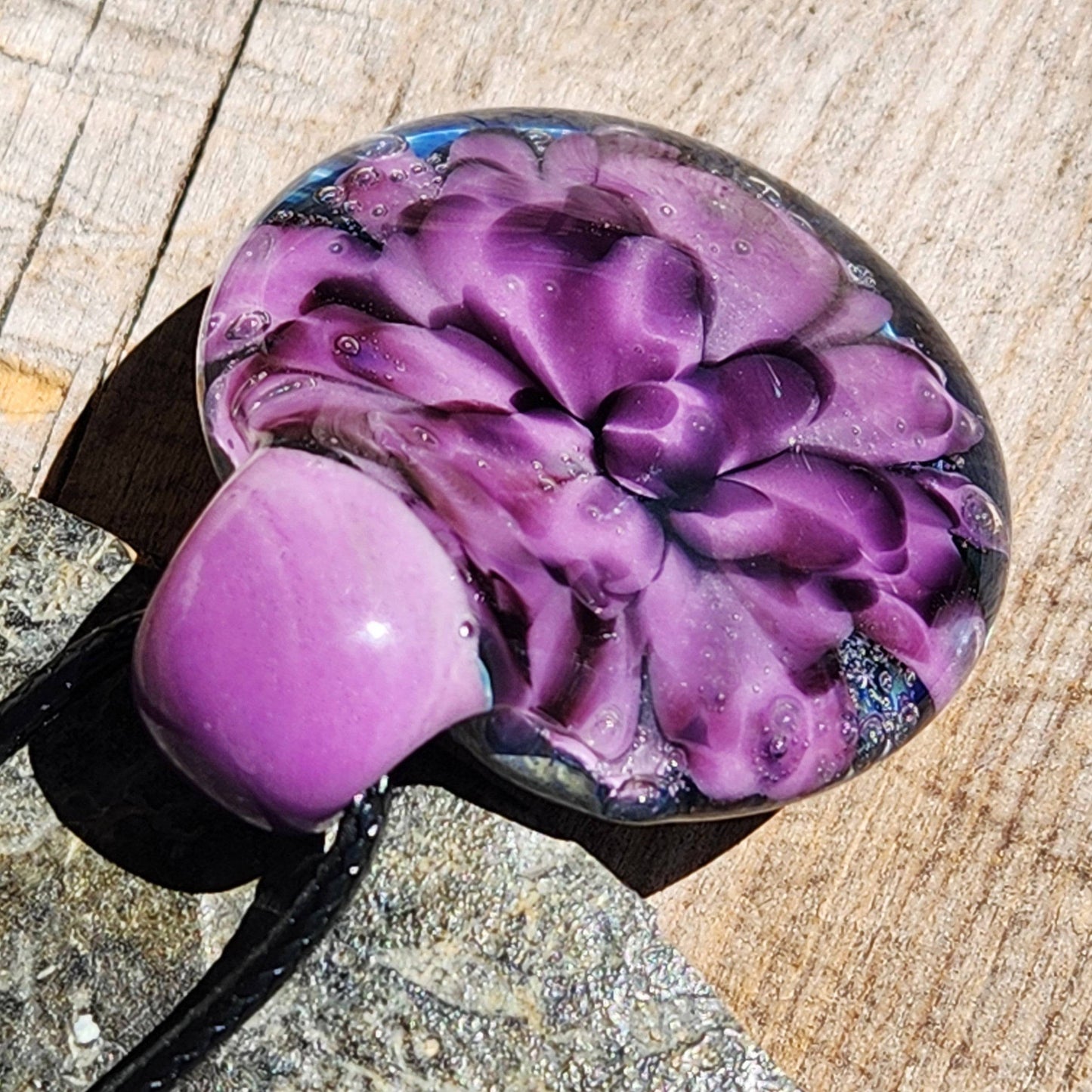 Artistic glass pendant with blooming purple flower design, resting on wood. Handcrafted borosilicate jewelry for unique style statements.