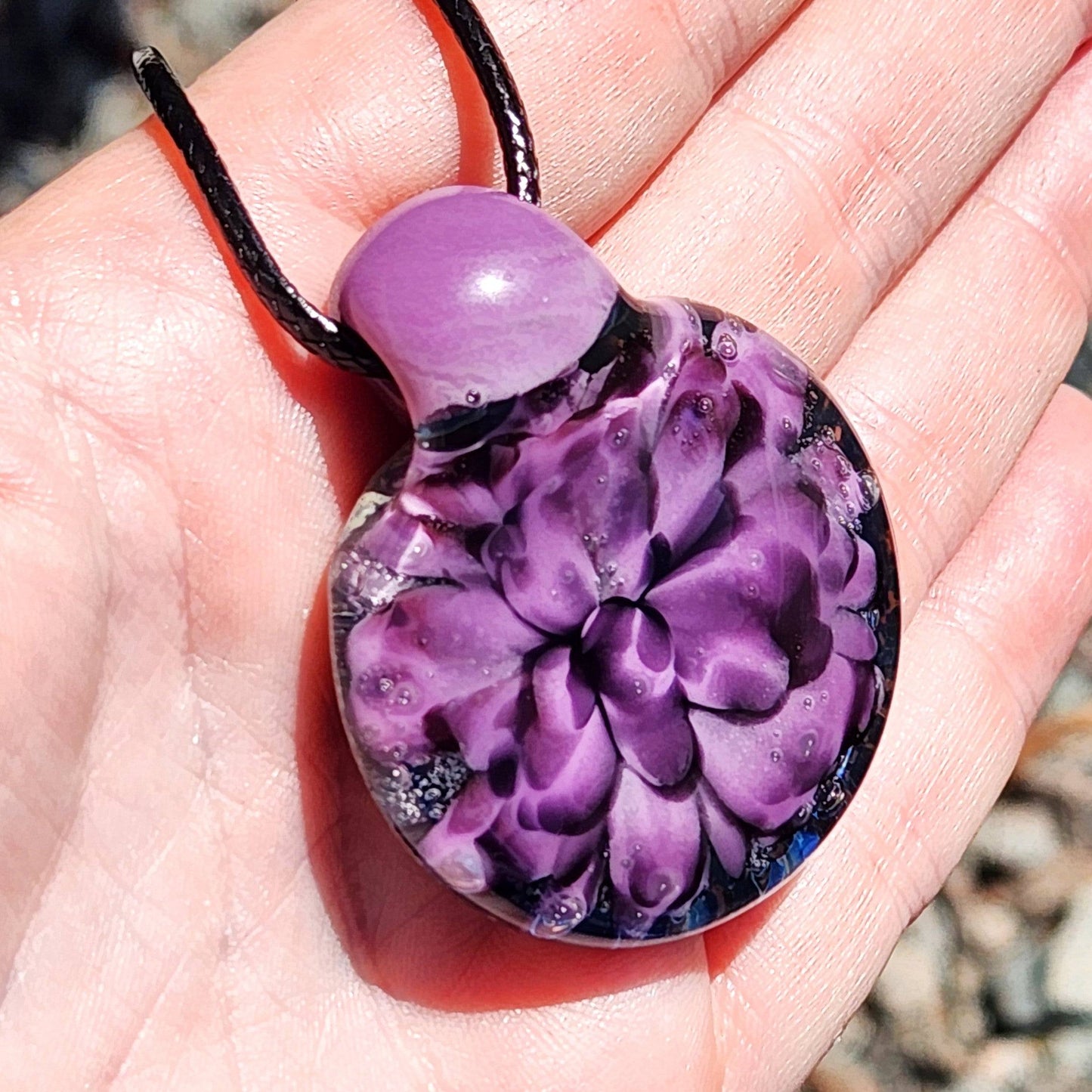 Close-up in hand of handmade glass pendant with purple flower encased in clear glass. Artistic and one-of-a-kind jewelry charm.