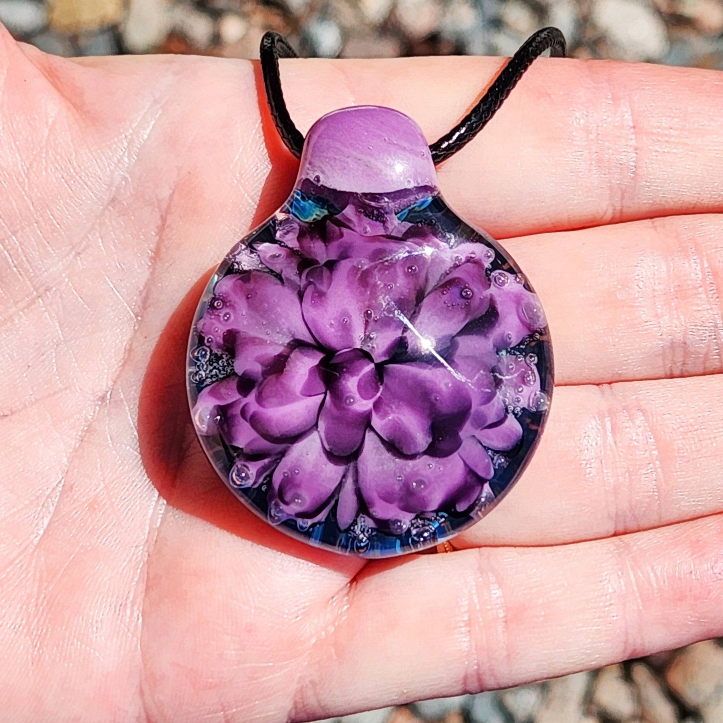 Hand holding a purple and black floral glass pendant necklace, showing size and detail. Unique borosilicate jewelry for everyday wear.