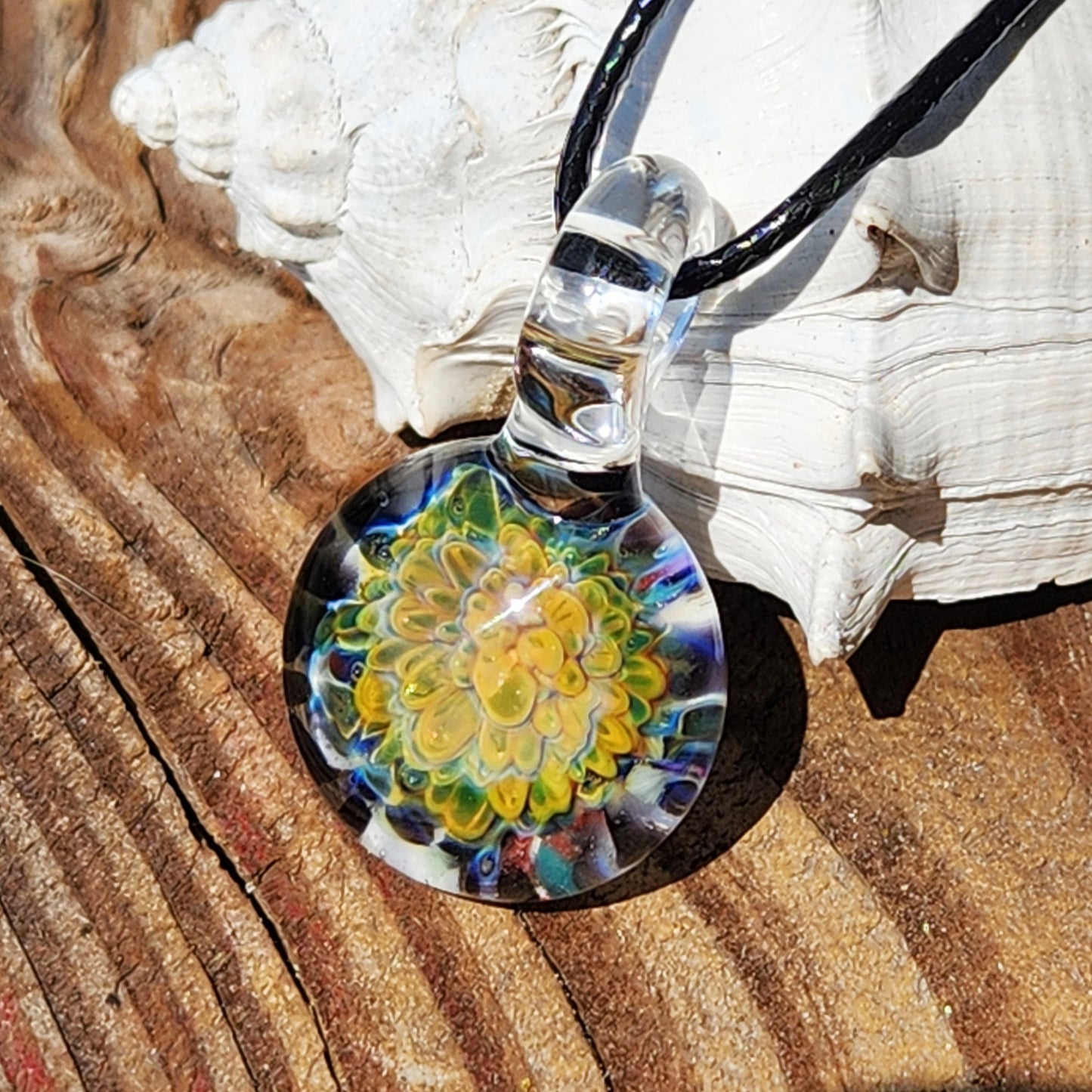 Handcrafted glass pendant with detailed multicolor flower, shown on a black cord necklace against a rustic wood background.