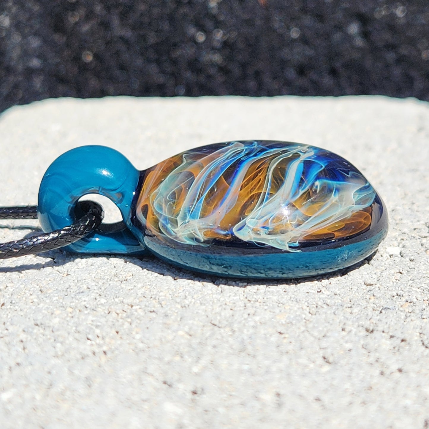 Side view of handmade blown glass pendant necklace showing layered gold and silver swirls. Borosilicate jewelry gift with blue glass loop and cord.