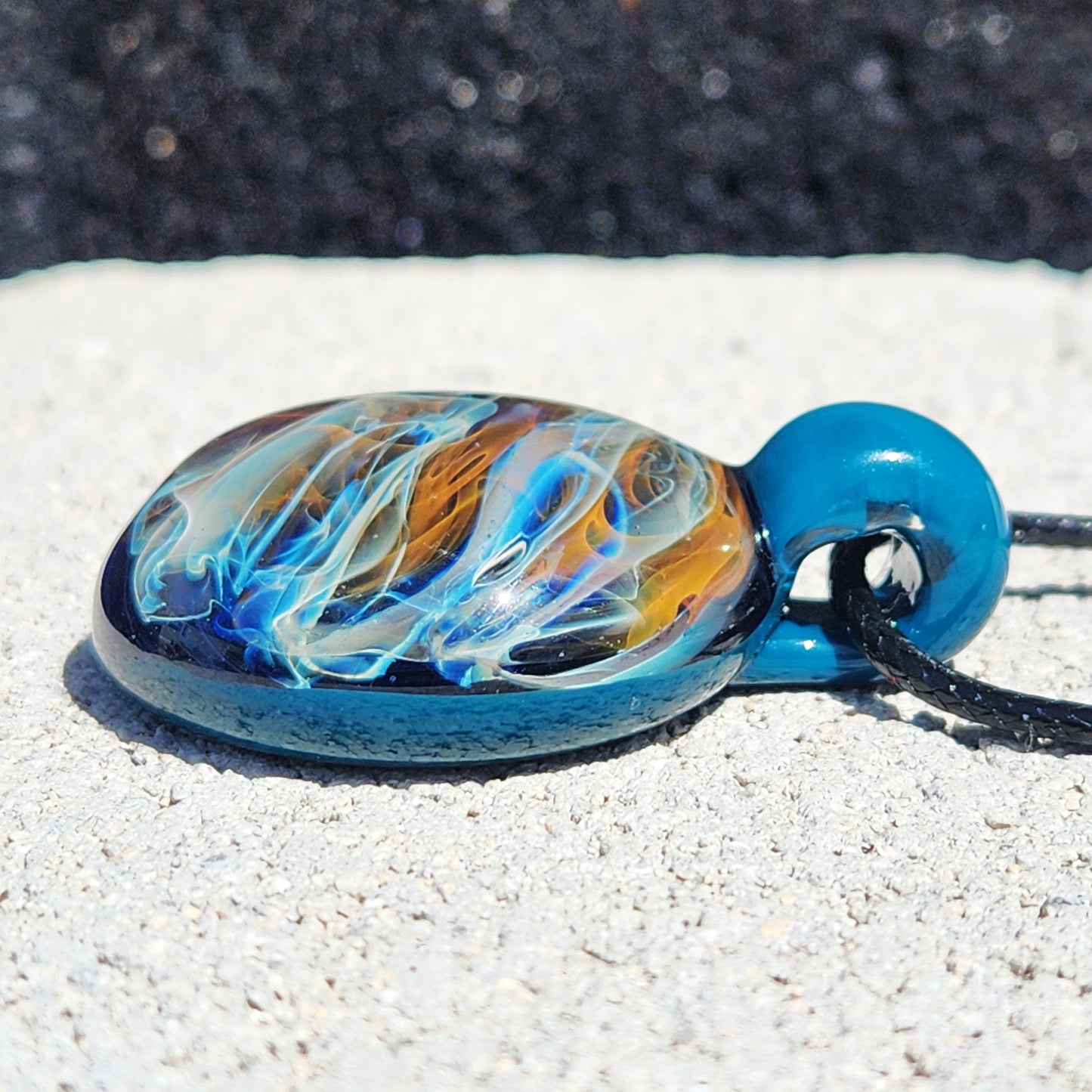 Side angle of handmade borosilicate glass pendant necklace with vibrant gold and silver swirls. Unique jewelry gift with blue loop and black cord.