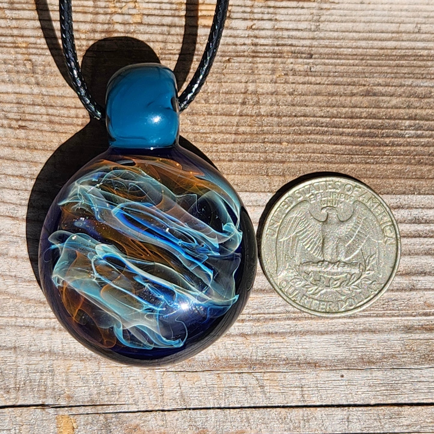 Handmade glass pendant necklace beside a US quarter for size reference. Borosilicate jewelry gift with swirling gold and silver colors on black cord.