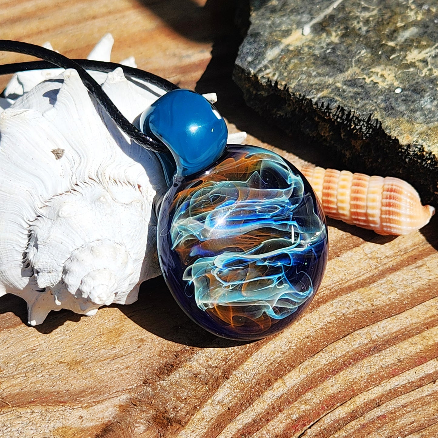 Handmade blown glass pendant with galaxy-like swirls in gold, silver, and blue. Borosilicate necklace gift displayed with seashells and natural textures.