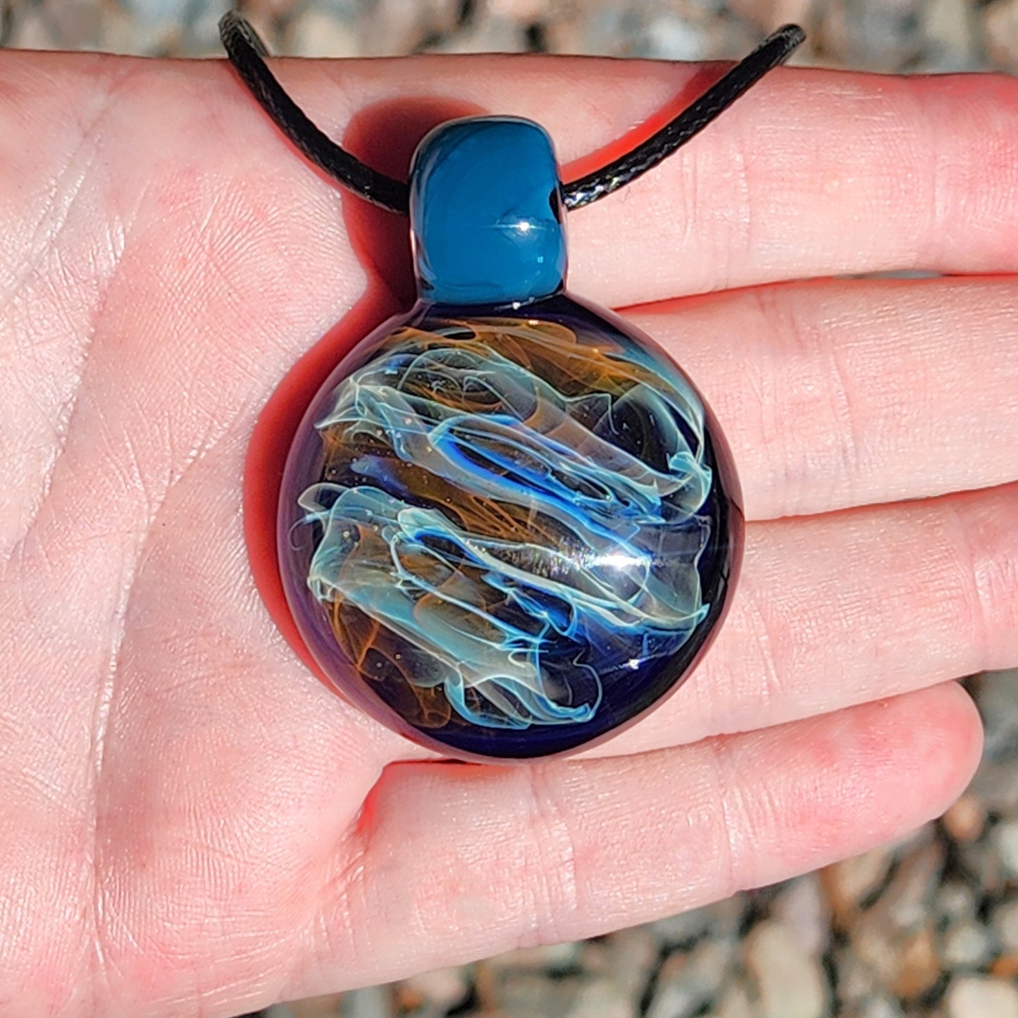 Front view of handmade glass pendant necklace in hand, showcasing vibrant gold and silver swirls. Borosilicate jewelry gift with blue glass loop.