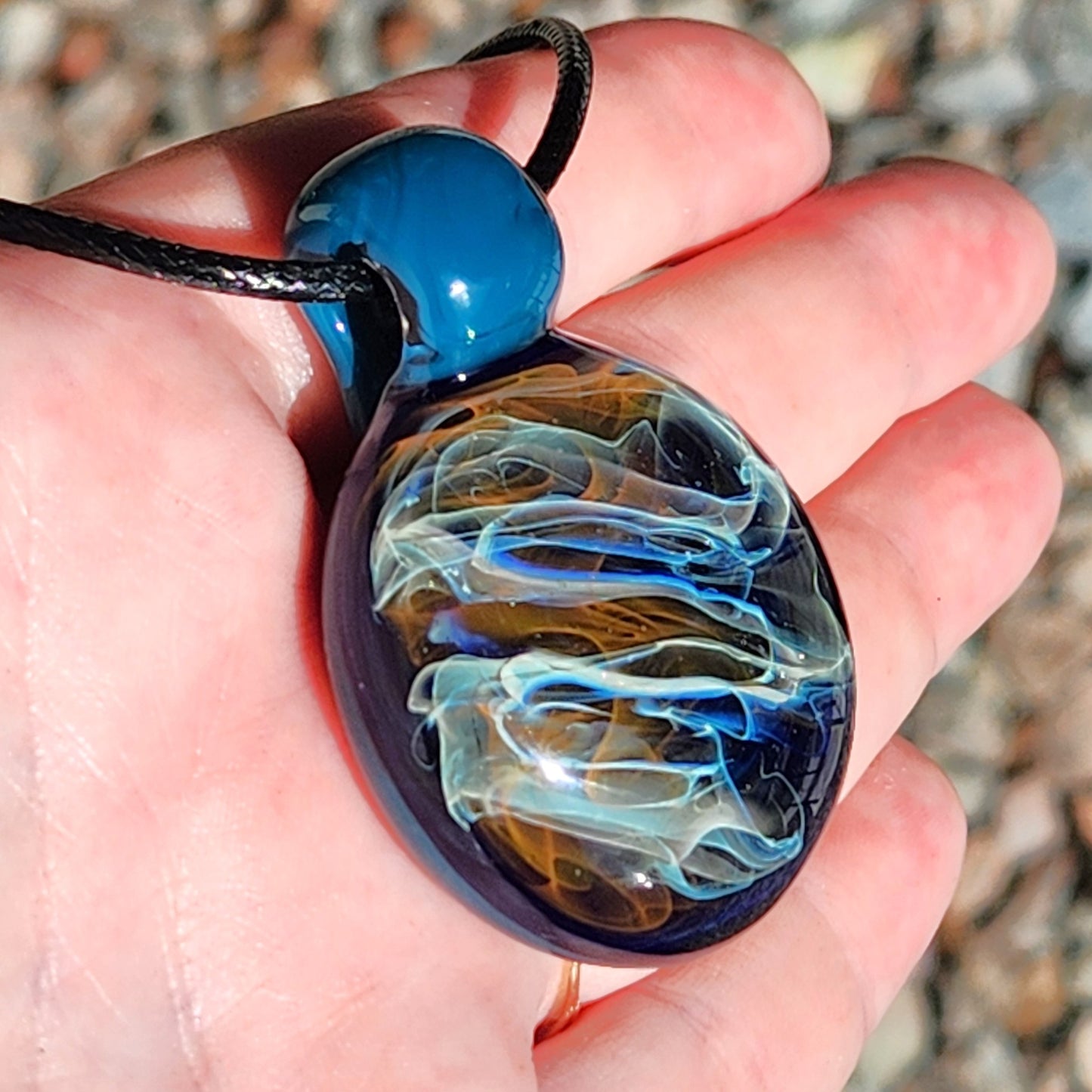 Hand holding handmade blown glass pendant necklace with swirling gold, silver, and blue hues. Unique borosilicate jewelry gift on adjustable cord.
