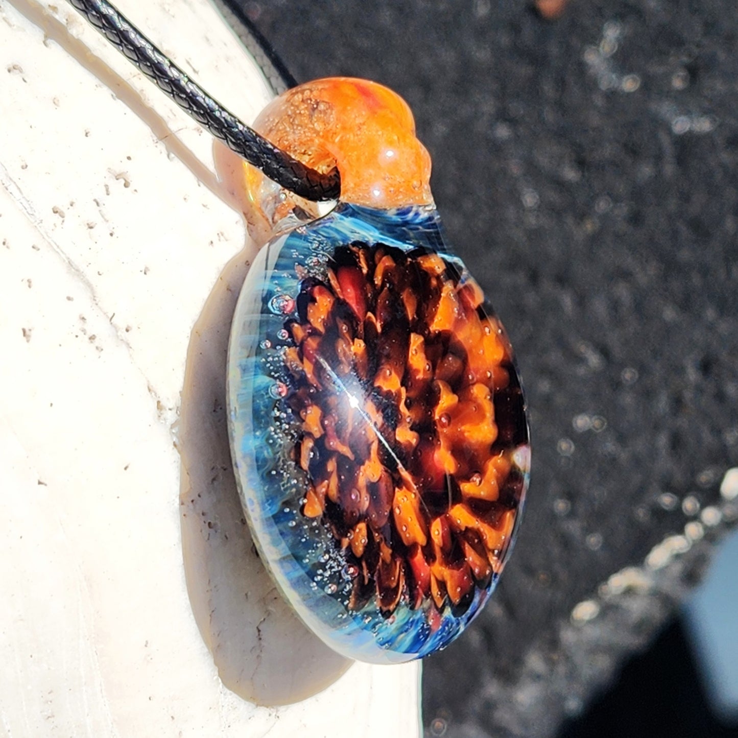Close-up of handmade borosilicate glass pendant with orange and red crushed glass, hanging on black cord. Artistic jewelry for gifting.