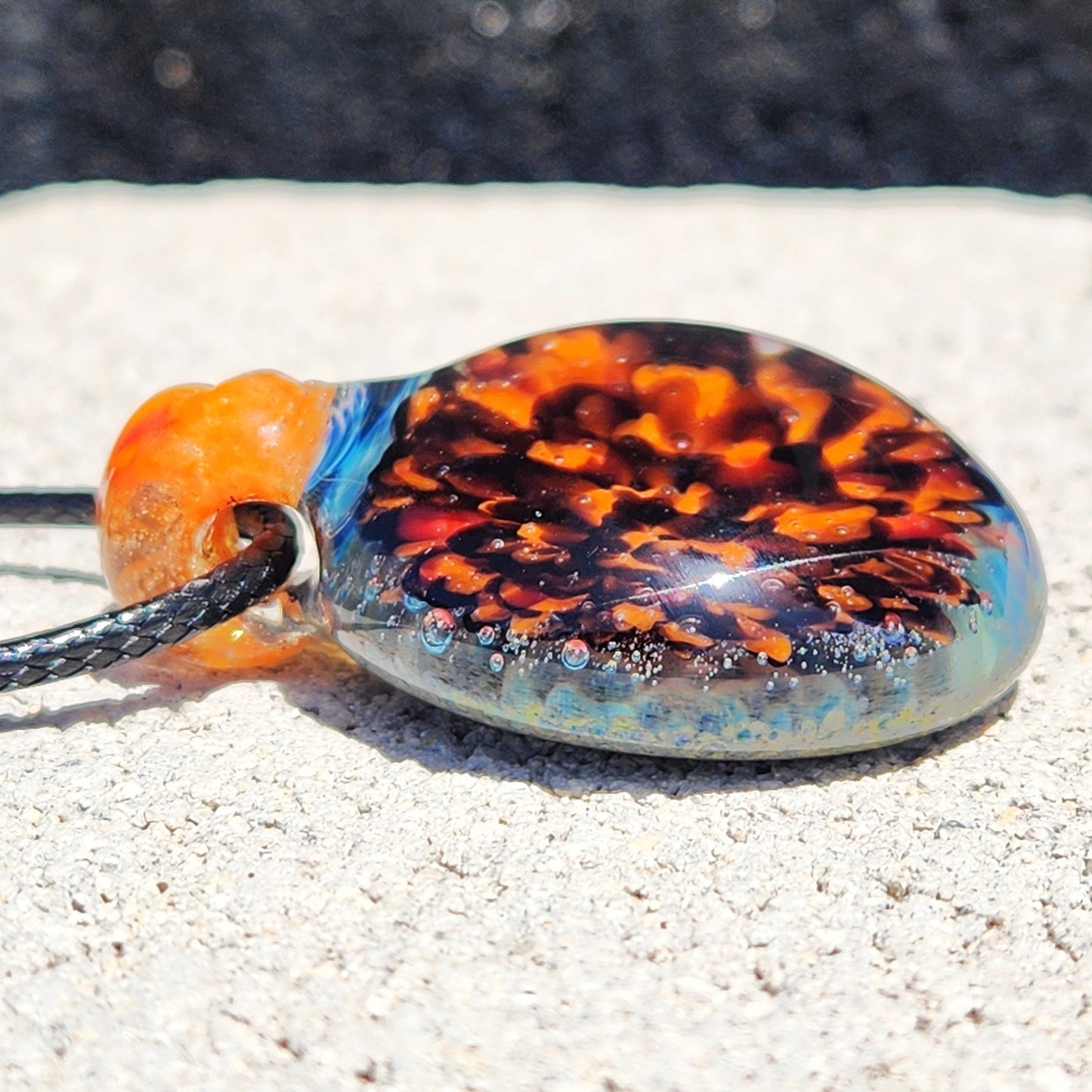 Side view of handmade glass pendant with fiery crushed borosilicate design, resting on stone. Unique necklace for apparel and accessories.