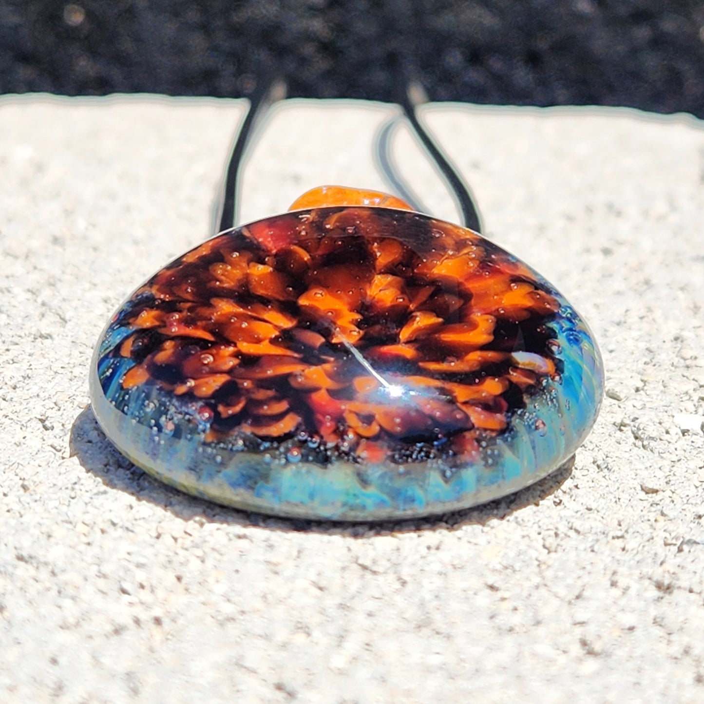 Crushed borosilicate glass pendant necklace in vivid orange and blue tones, lying flat on stone. Handmade jewelry for apparel accessories.