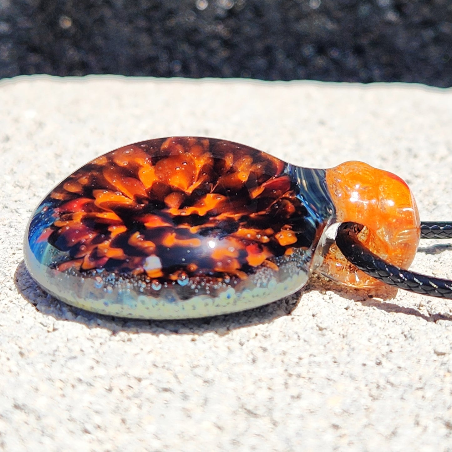 Handblown glass pendant with vibrant orange crushed borosilicate, lying on textured surface. Handmade necklace for jewelry enthusiasts.