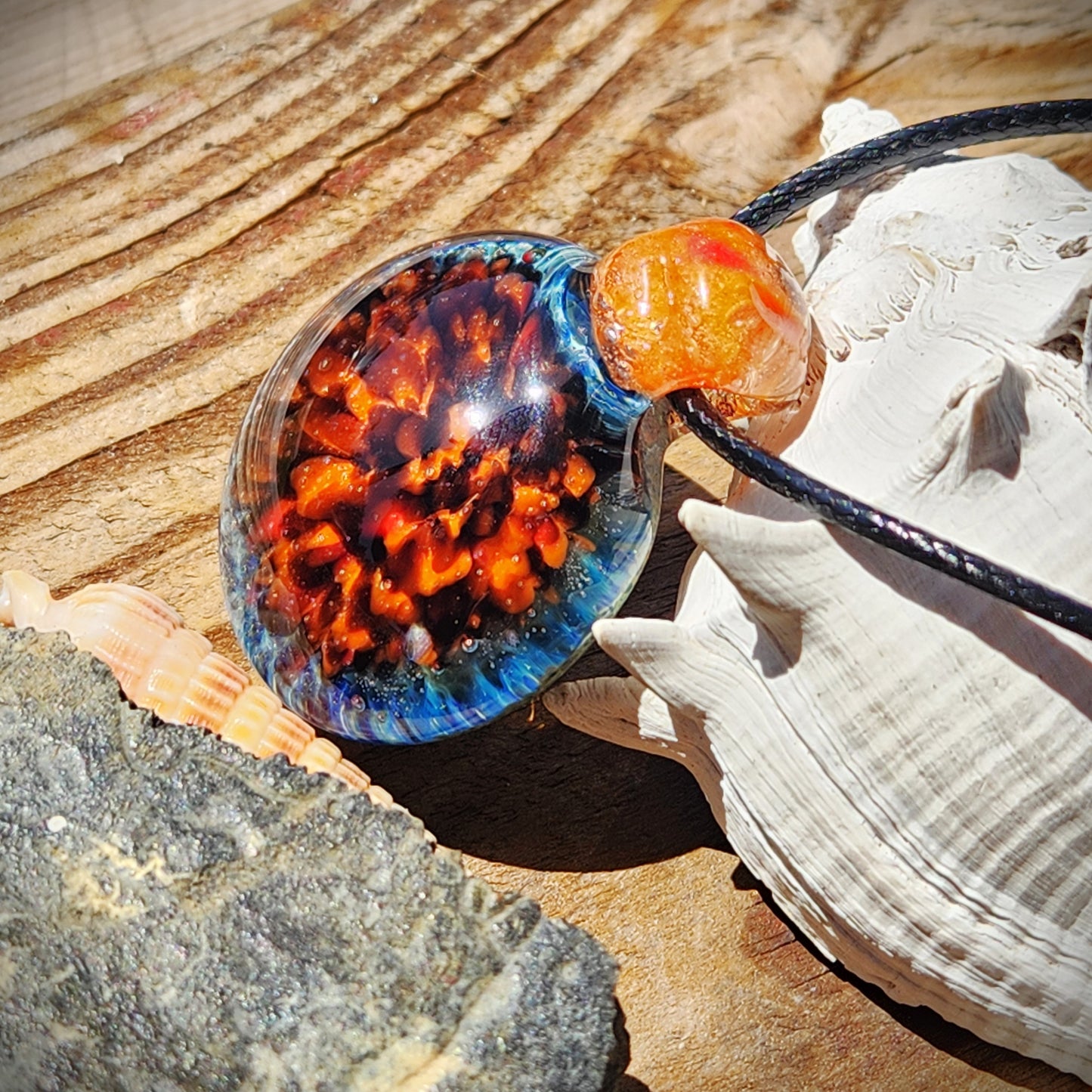 Colorful handmade glass pendant with crushed borosilicate core, displayed with seashells and rocks. Unique gift-ready necklace for women.