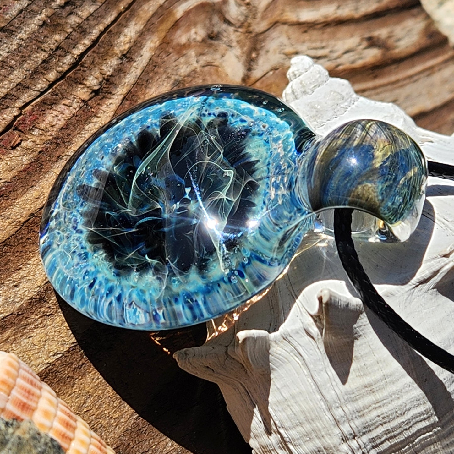 Handmade Pendant Necklace – Winter Scene Borosilicate Glass Pendant on wood, glowing in sunlight with vivid blue and silver swirls.