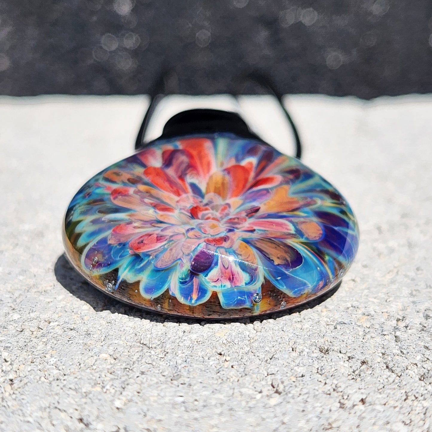 Close-up of handmade glass pendant showing detailed lampworked flower in red, orange, and blue hues. Artisan jewelry gift for her.