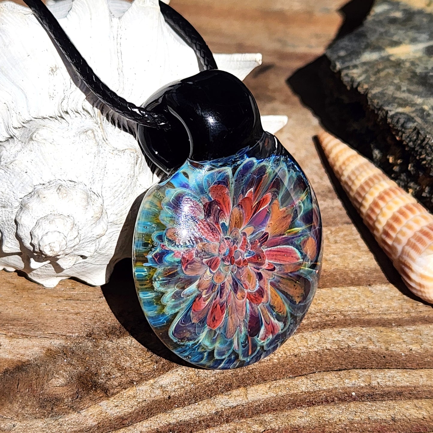 Artisan blown glass pendant necklace resting on wood with seashells. Features colorful floral design in borosilicate glass.