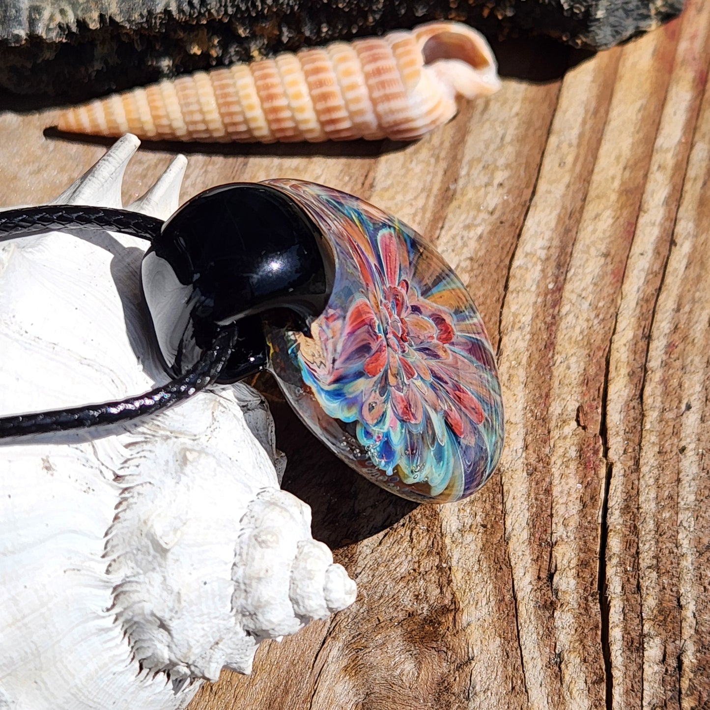 Angled view of handmade glass pendant on shell background. Features colorful flower design in borosilicate glass.
