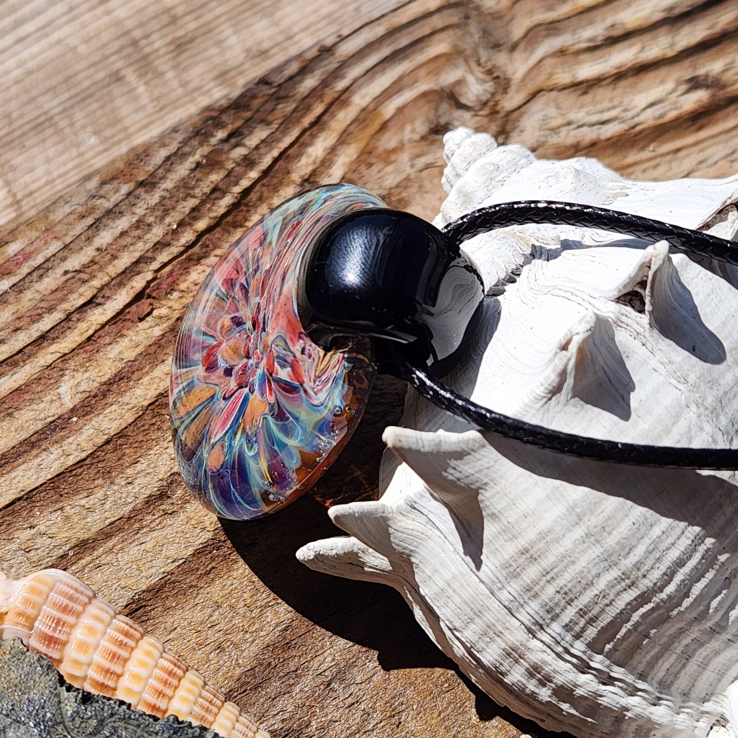 Side view of artisan glass pendant necklace on wood with seashells. Handmade jewelry gift with colorful floral design.