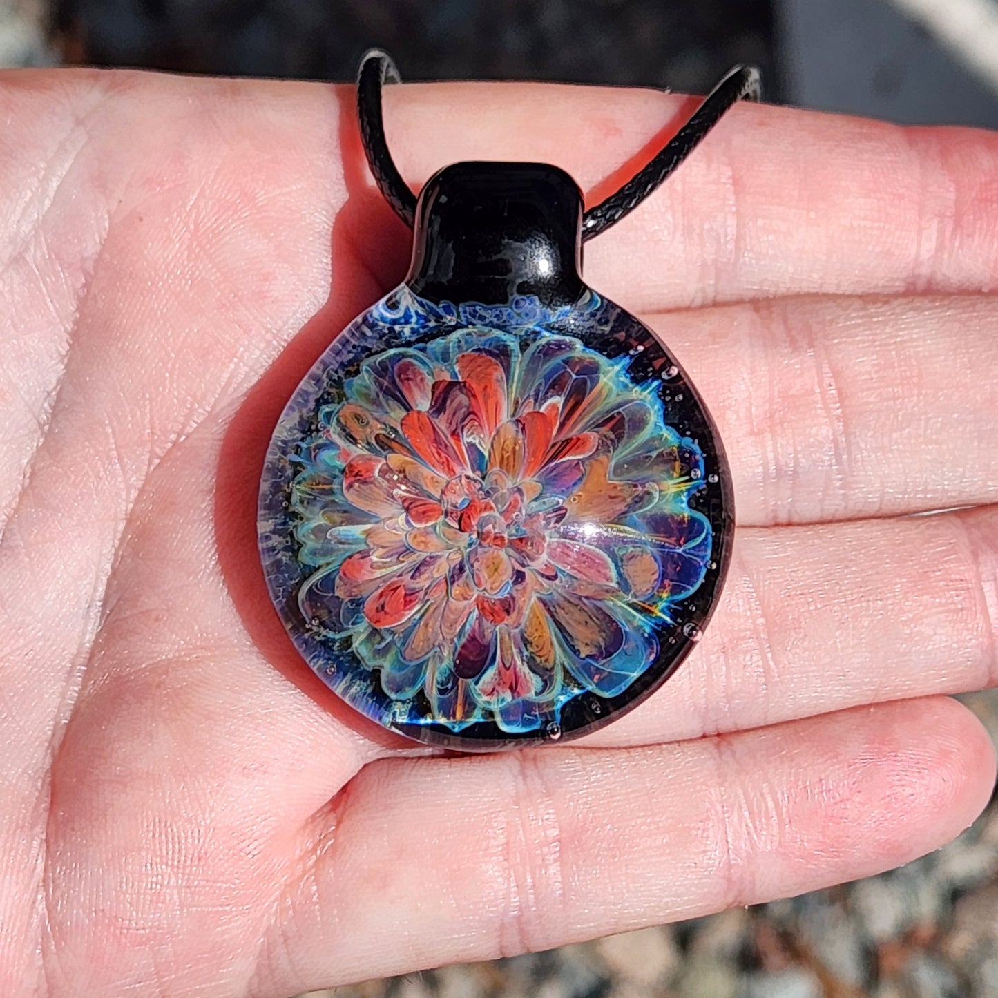 Hand holding handmade blown glass pendant necklace. Features red, orange, and blue flower design in borosilicate glass.
