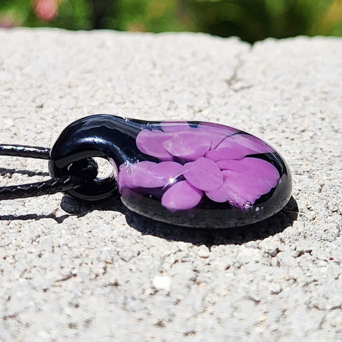 Side view of small handmade purple flower glass pendant on black cord, highlighting its smooth, domed surface and vibrant color.