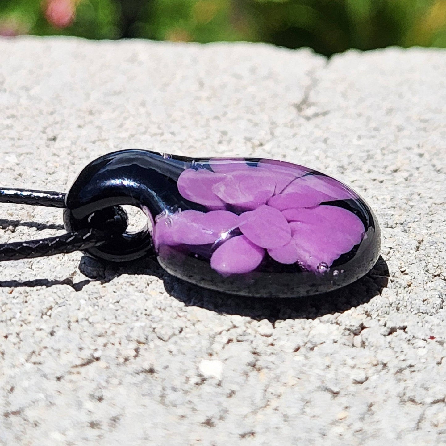 Side view of small handmade purple flower glass pendant on black cord, highlighting its smooth, domed surface and vibrant color.