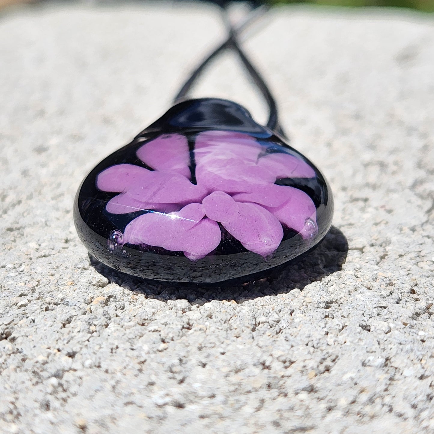 Close-up of small handmade purple flower glass pendant on black cord, showcasing glossy finish and intricate petal details.