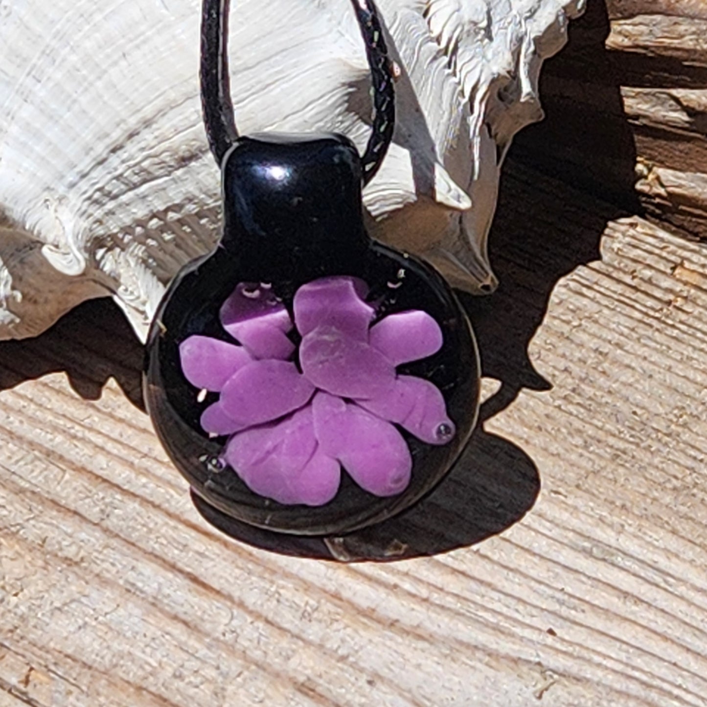 Sunlit handmade purple flower glass pendant on black cord, perfect as a lightweight, artistic gift for him or her.