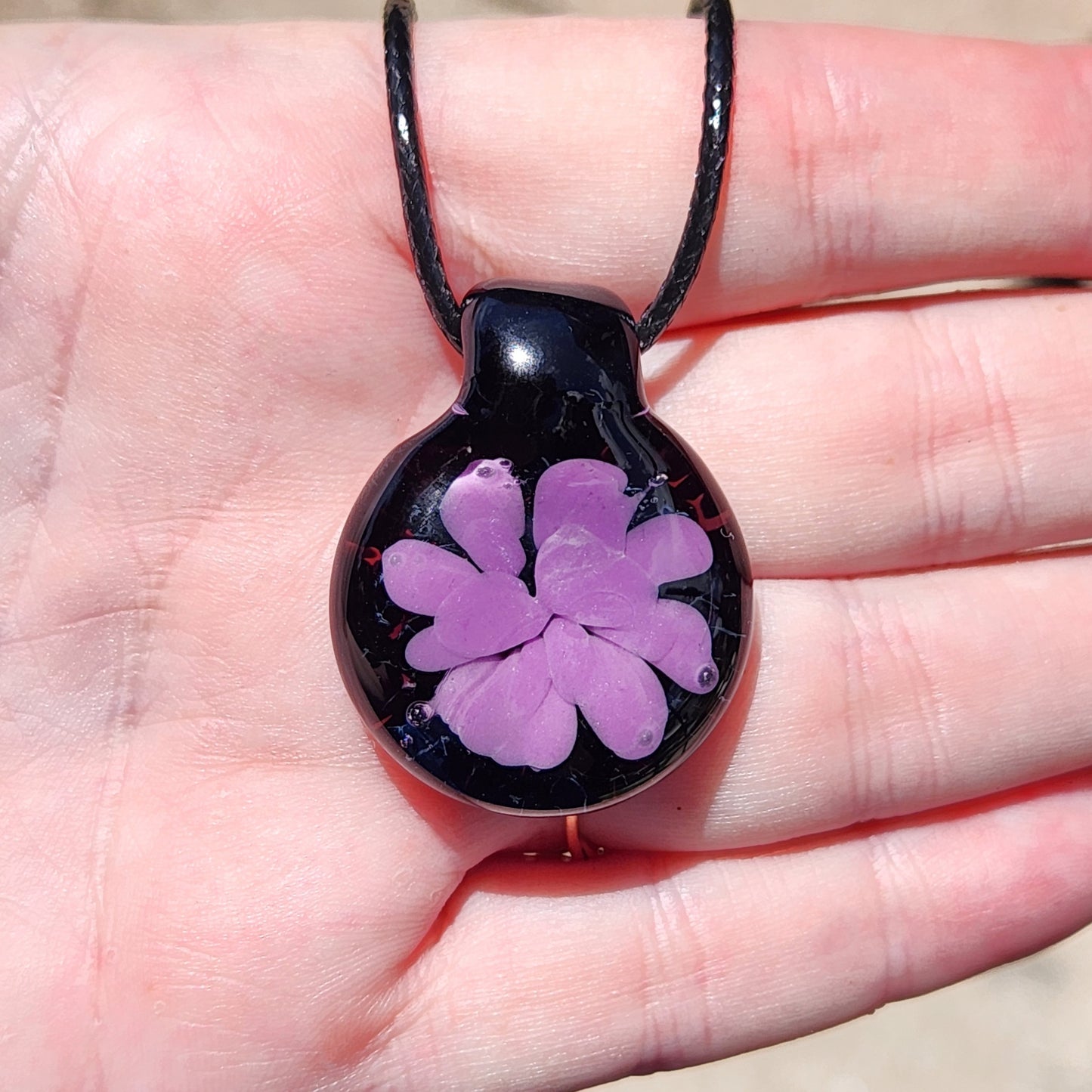 Handheld view of minimalist purple flower glass pendant on black cord, showcasing its size and elegant handmade detail.