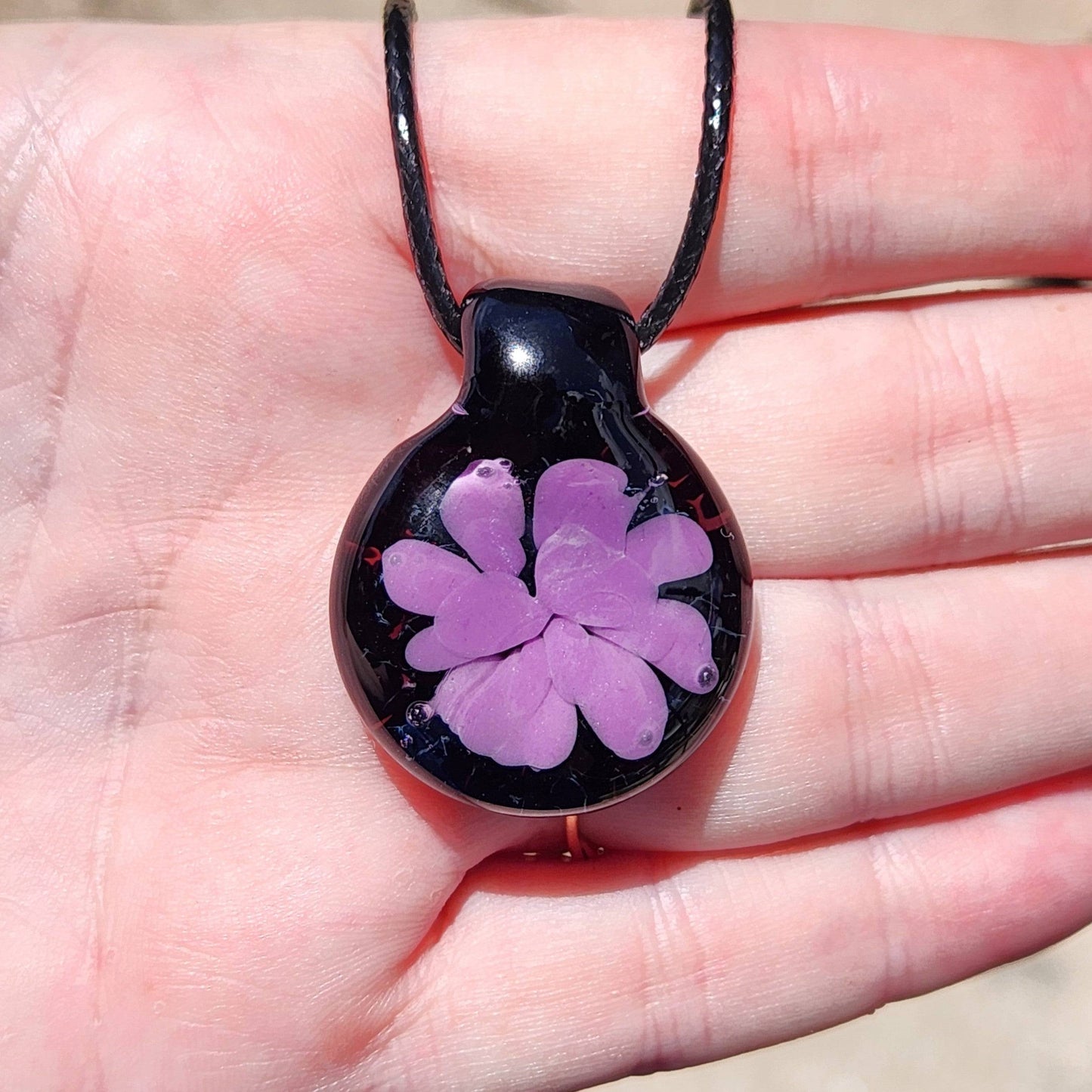 Handheld view of minimalist purple flower glass pendant on black cord, showcasing its size and elegant handmade detail.