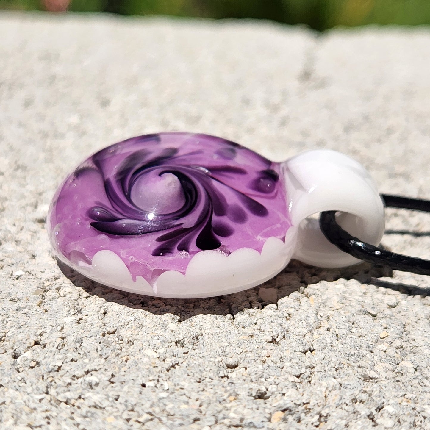 Close-up of handmade purple spiral glass pendant necklace with white and black detailing, lying flat on a stone surface.