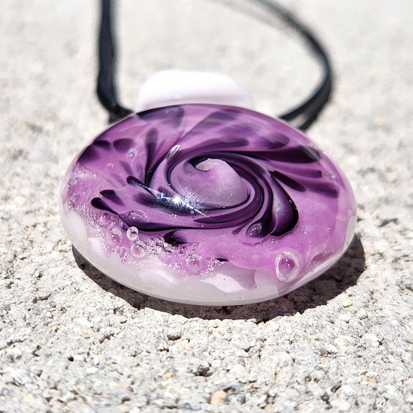 Close-up of handmade purple spiral glass pendant necklace with black accents and white base, shown on a concrete background.