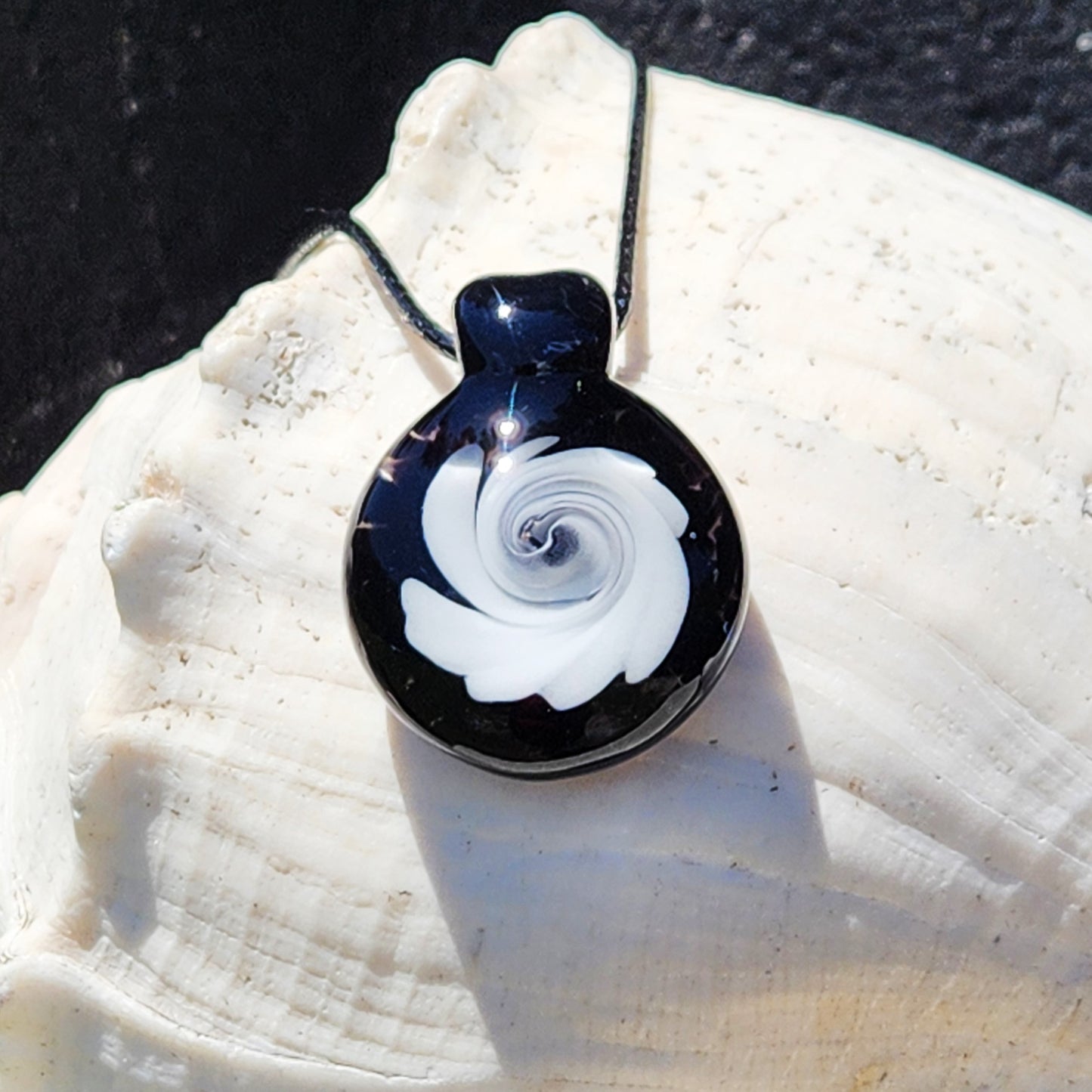 Handmade black and white swirl glass pendant necklace resting on a white seashell, showcasing its elegant spiral pattern and polished surface.