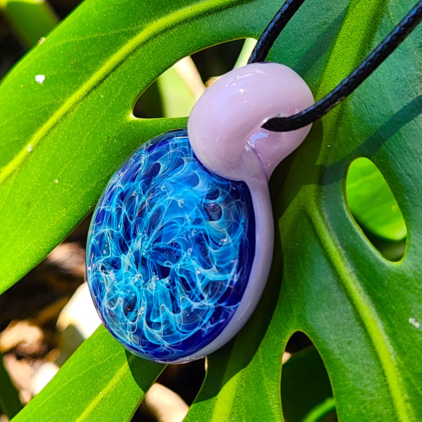 Heady glass pendant necklace with vivid blue swirls and pink loop resting on green leaf. Handmade charm with dreamy silver fuming.
