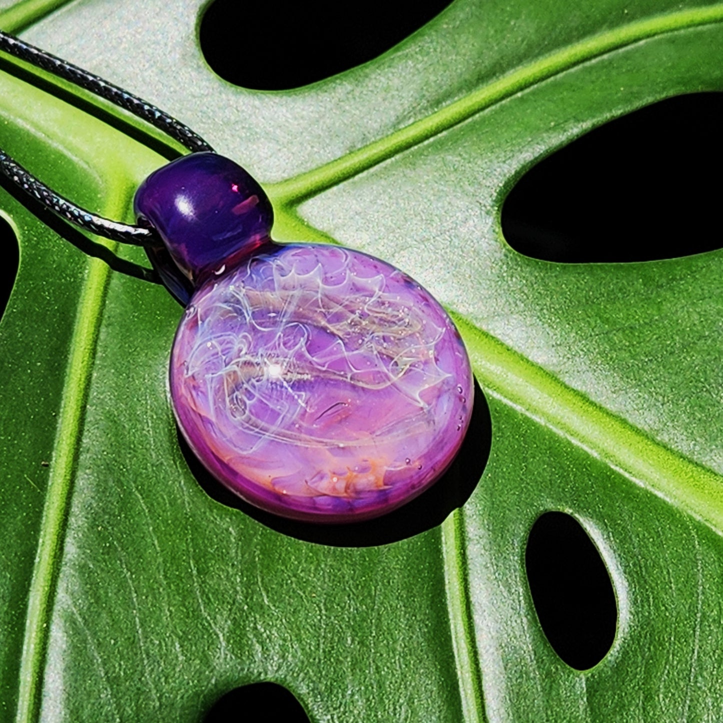 Purple glass pendant necklace with silver and gold fuming displayed on a green monstera leaf. Vibrant artisan jewelry for nature lovers.