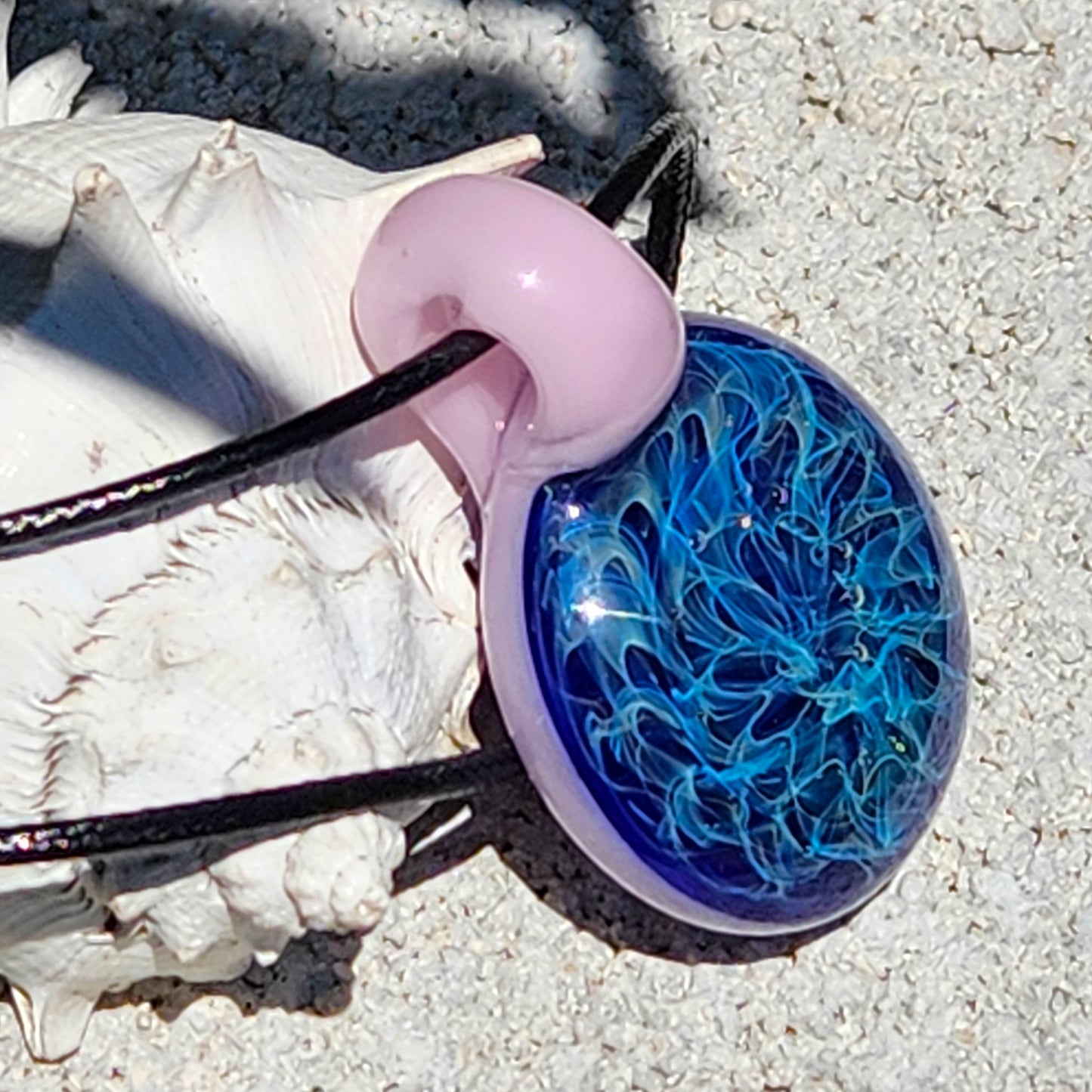Close-up of blue and pink handmade glass pendant necklace on sand. Swirling design with silver fuming for a dreamy accessory.